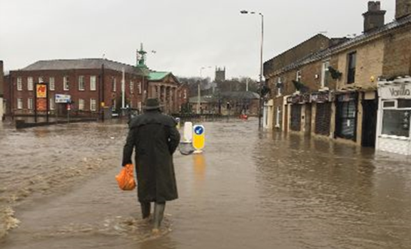 Man with green wellington boots on walks through flood waters carrying a orange plastic bag. Padiham Town Hall is in the background. 