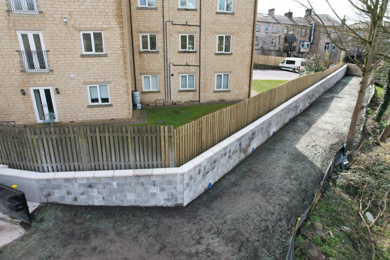 New grey wall along the riverbank with wooden fence panels behind and a block of flats
